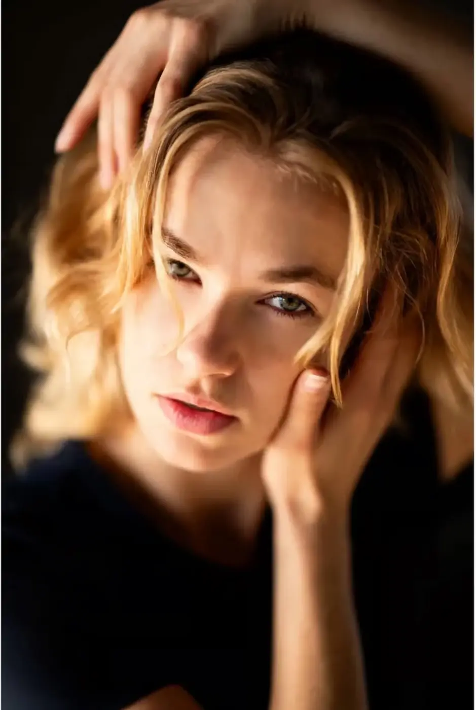 Close-up portrait of a blonde woman with her hands around her face and a strongly blurred background.