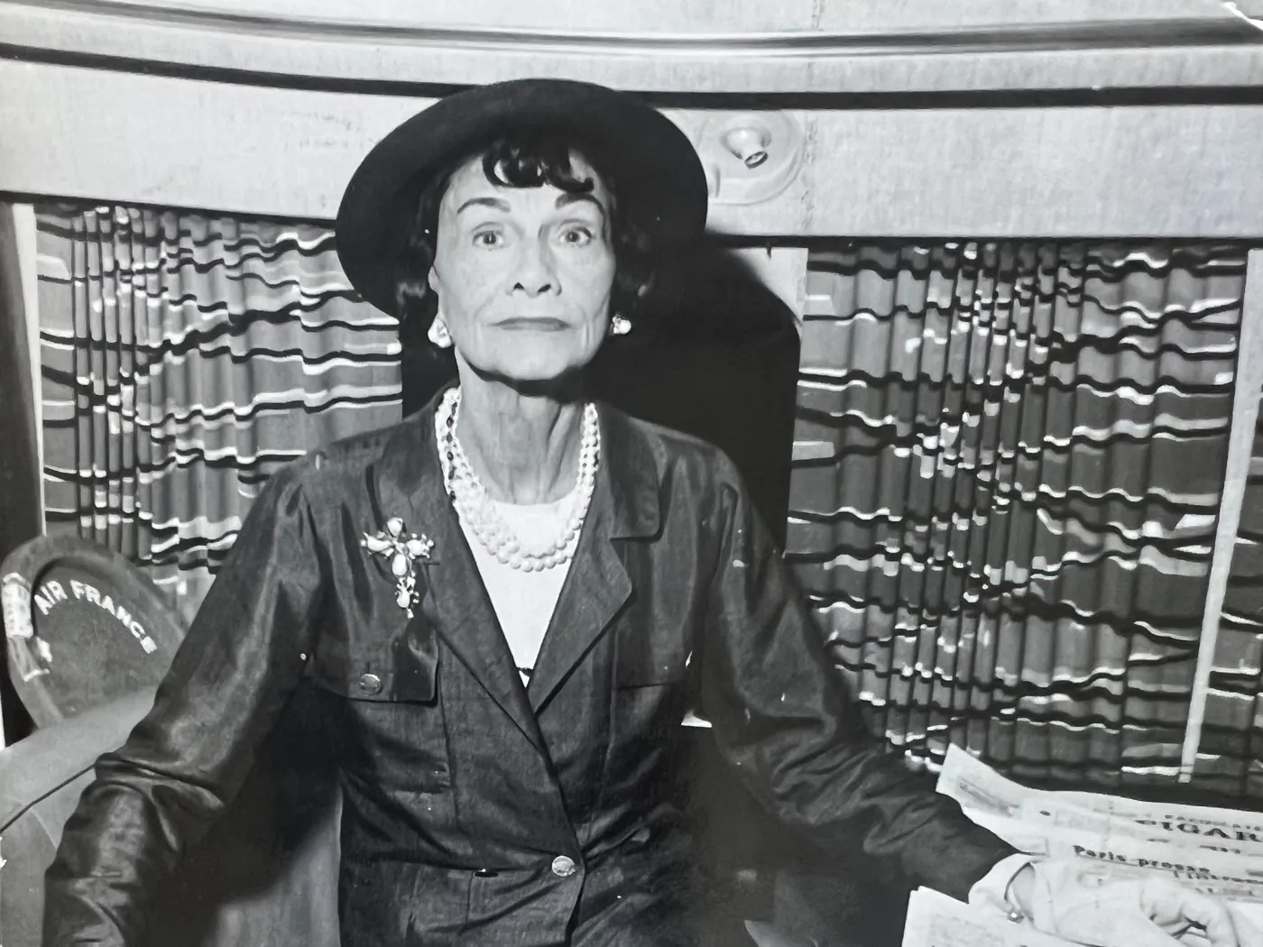 Zwart-witfoto van Coco Chanel met hoed en parelkettingen, zittend naast een stapel kranten.
