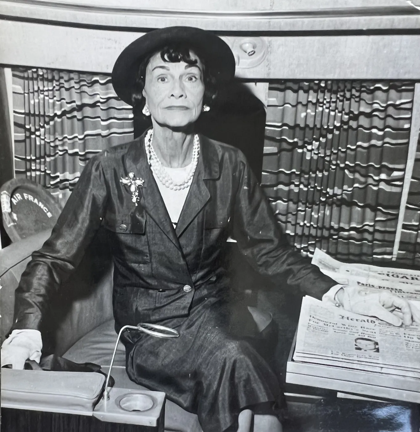 Black-and-white photograph of Coco Chanel wearing a hat and pearl necklaces, seated beside a stack of newspapers.