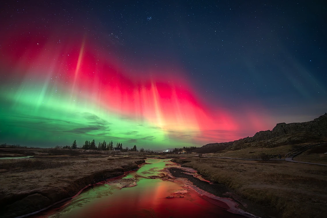 Rood en groen noorderlicht weerspiegelt in een waterloop tussen donkere oevers in Þingvellir op IJsland.