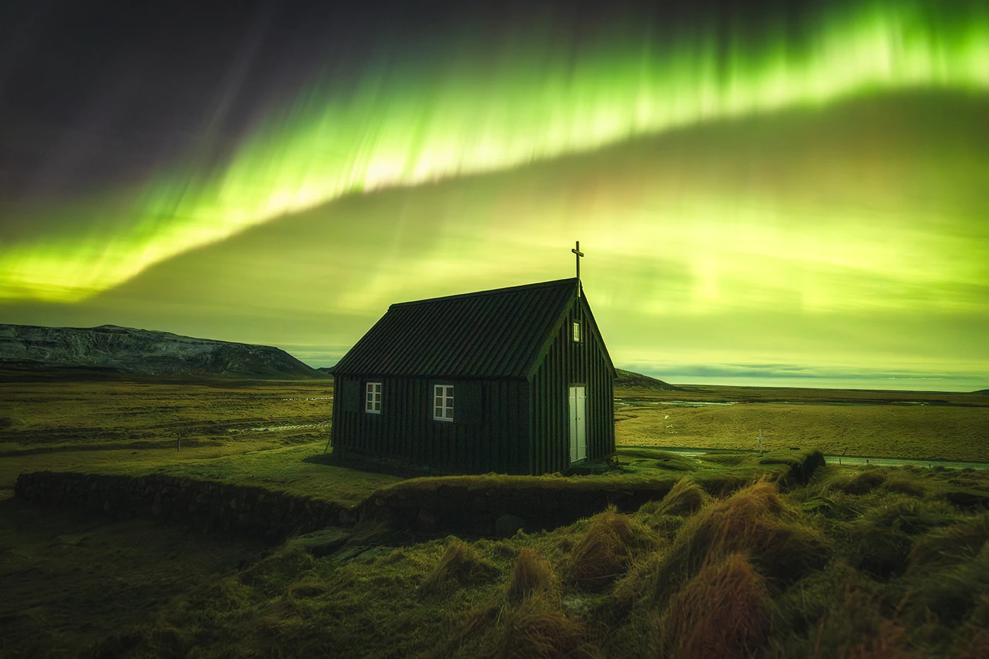 Groene banen noorderlicht boven een donker houten kerkje in een open IJslands landschap.