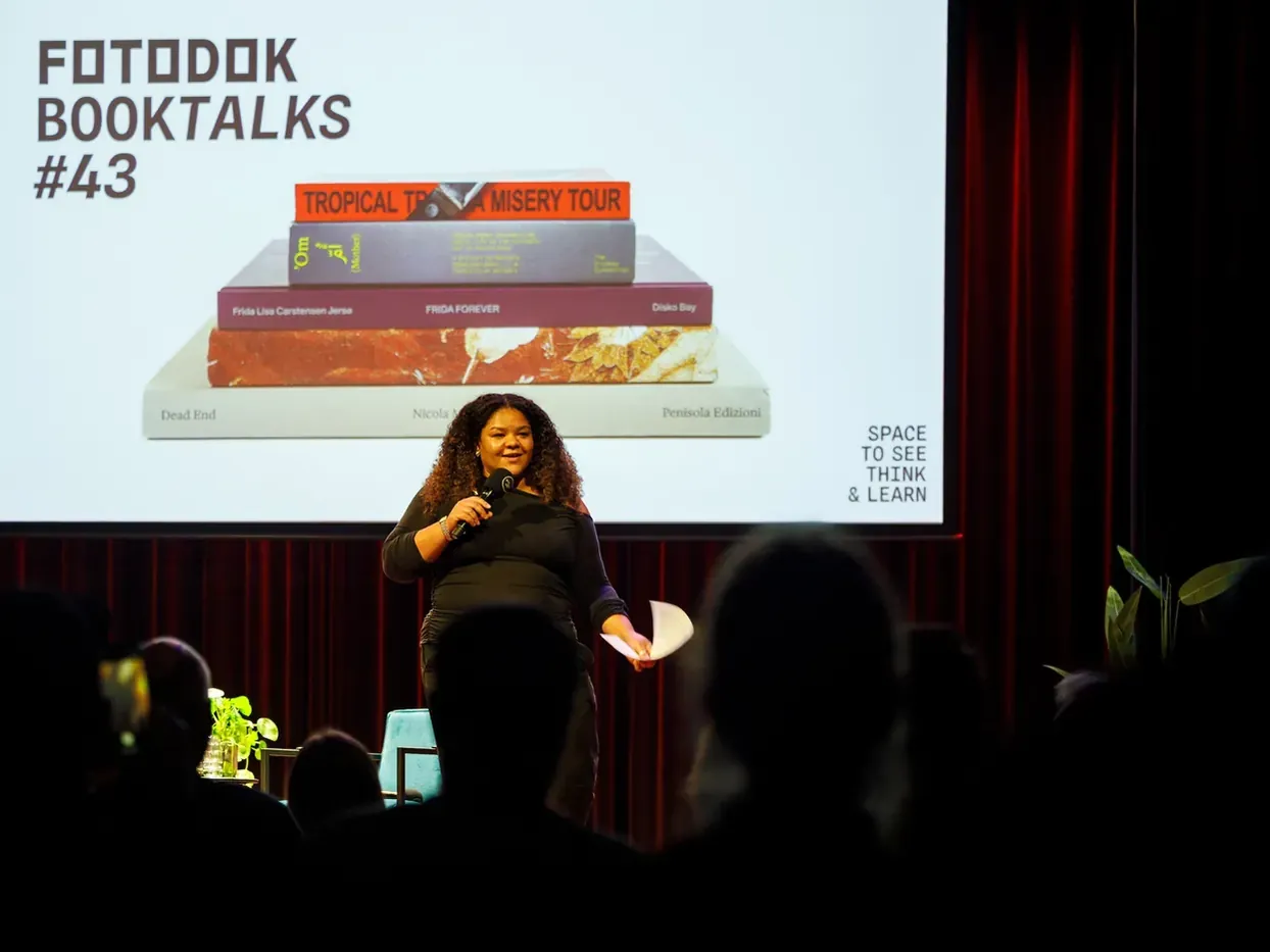 Presentator Giya Makondo-Wills spreekt op het podium tijdens FOTODOK BOOK TALKS #43, met fotoboeken op het scherm achter haar.