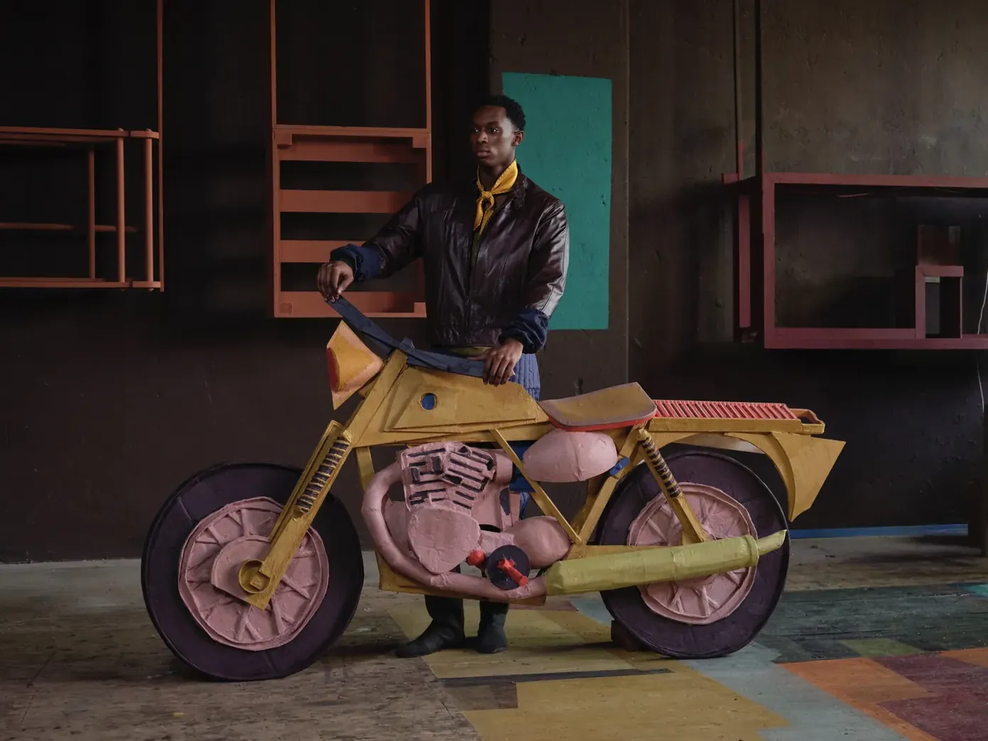 Een persoon met een gele halsdoek houdt een grote geel-roze motorfiets van textiel voor zich in een ruimte met gekleurde vlakken.