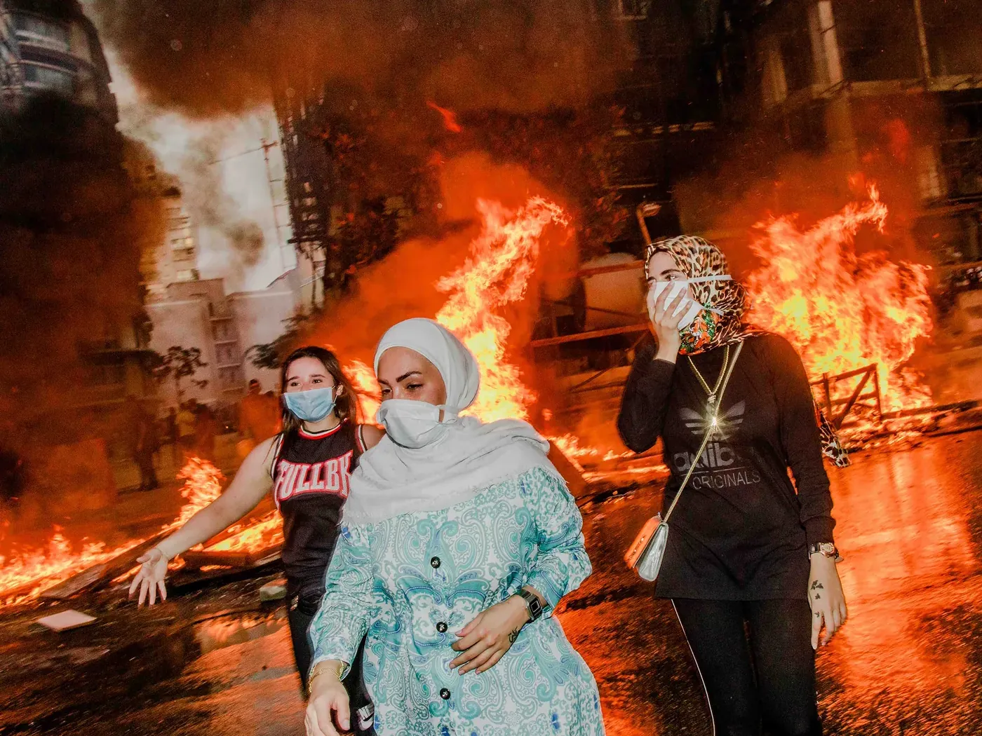 Drie gemaskerde vrouwen lopen langs grote vuren in een stadsstraat, gefotografeerd door Myriam Boulos.
