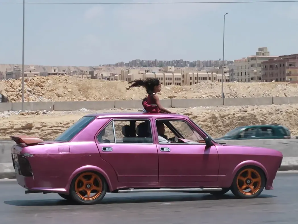 Een meisje staat door het open dak van een rijdende roze auto in Caïro, gefotografeerd door Yomna El Beyaly.
