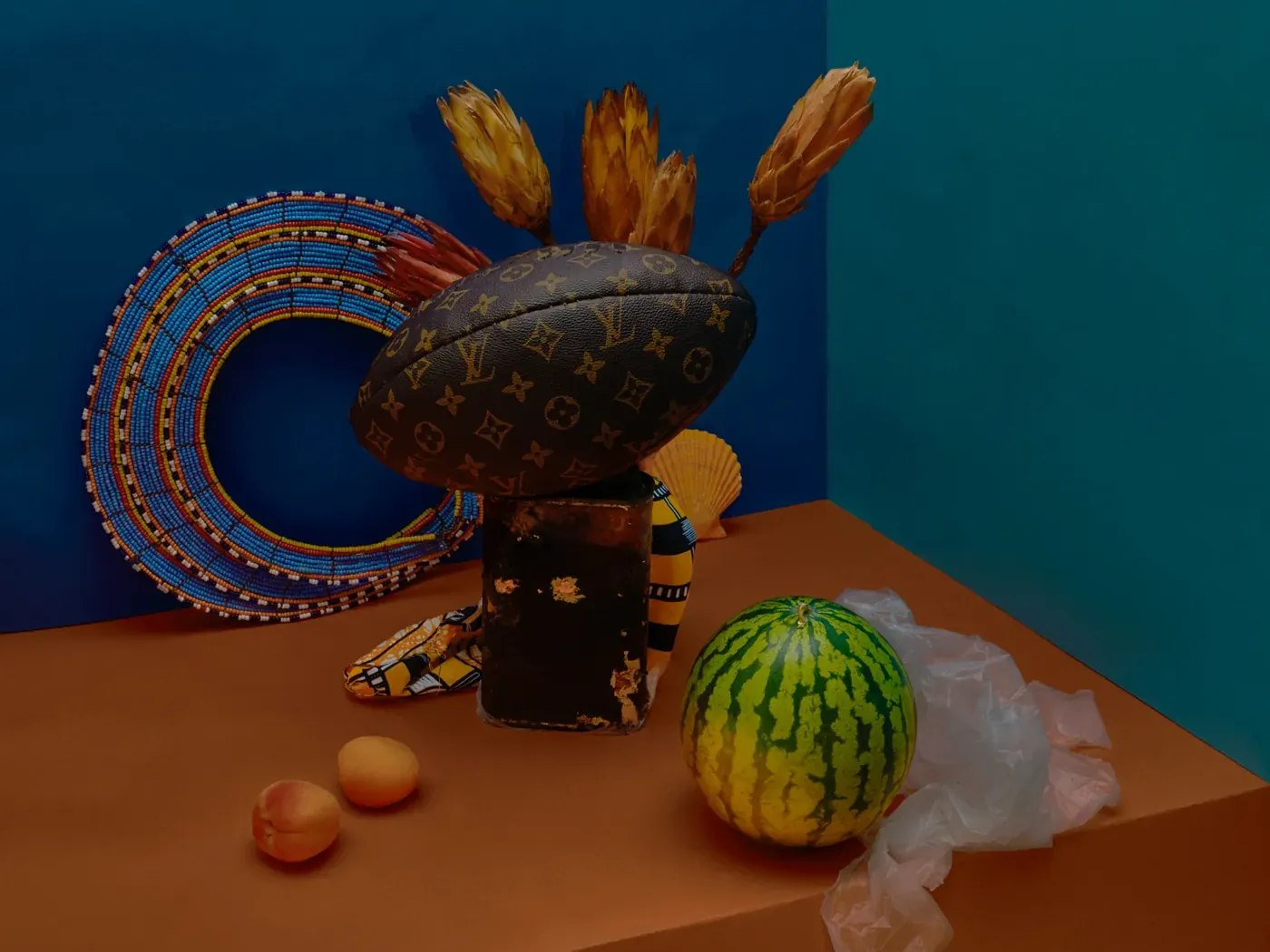 Fotografisch stilleven met een kralencirkel, een bedrukte etui, gedroogde bloemen en fruit tegen blauwe wanden van Maya Inès Touam.