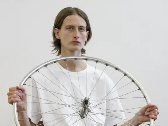 Een persoon in een wit T-shirt houdt zittend een fietswiel voor zich vast in werk van Andrzej Steinbach.