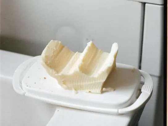 Een deels uitgeschept stuk boter met diepe groeven op een witte botervloot, naast een keukenwerkblad.