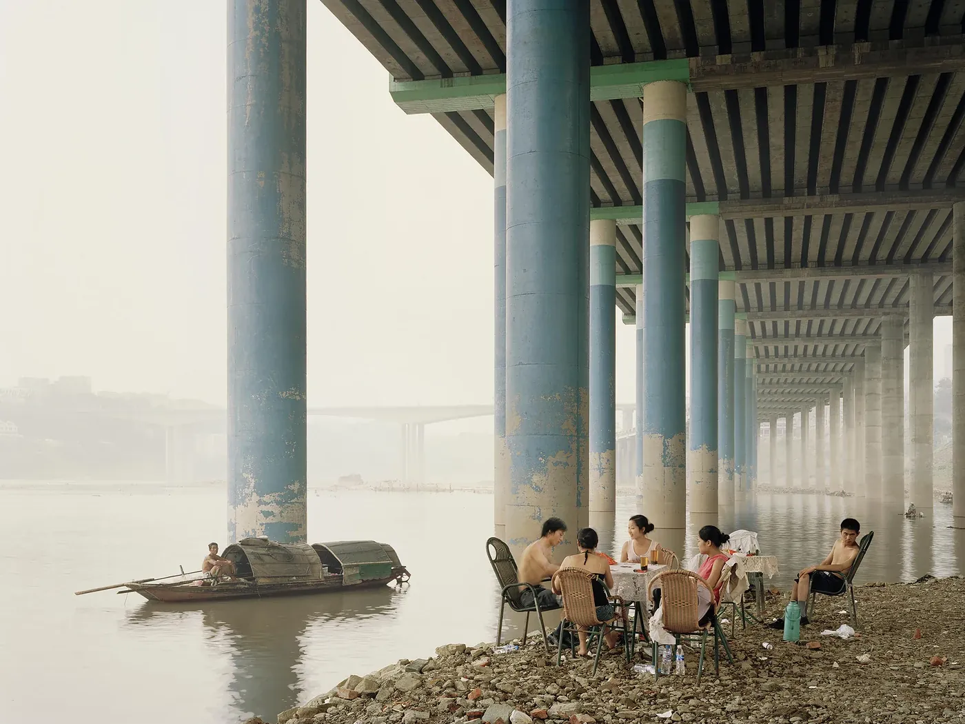 Mensen zitten aan een tafel onder een brug bij een rivier, terwijl een kleine boot voorbijvaart.