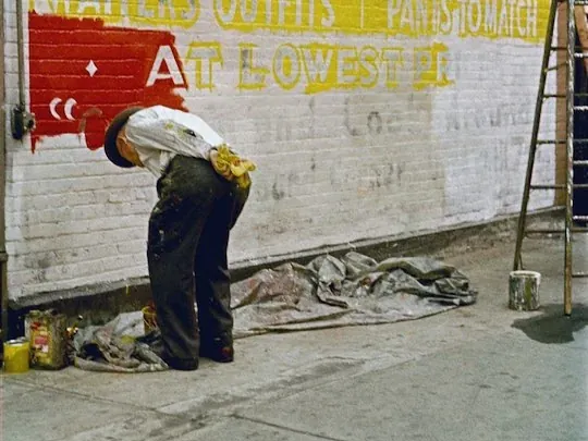 Een schilder werkt aan een gele en rode reclameschildering op een bakstenen muur, gefotografeerd door Saul Leiter.