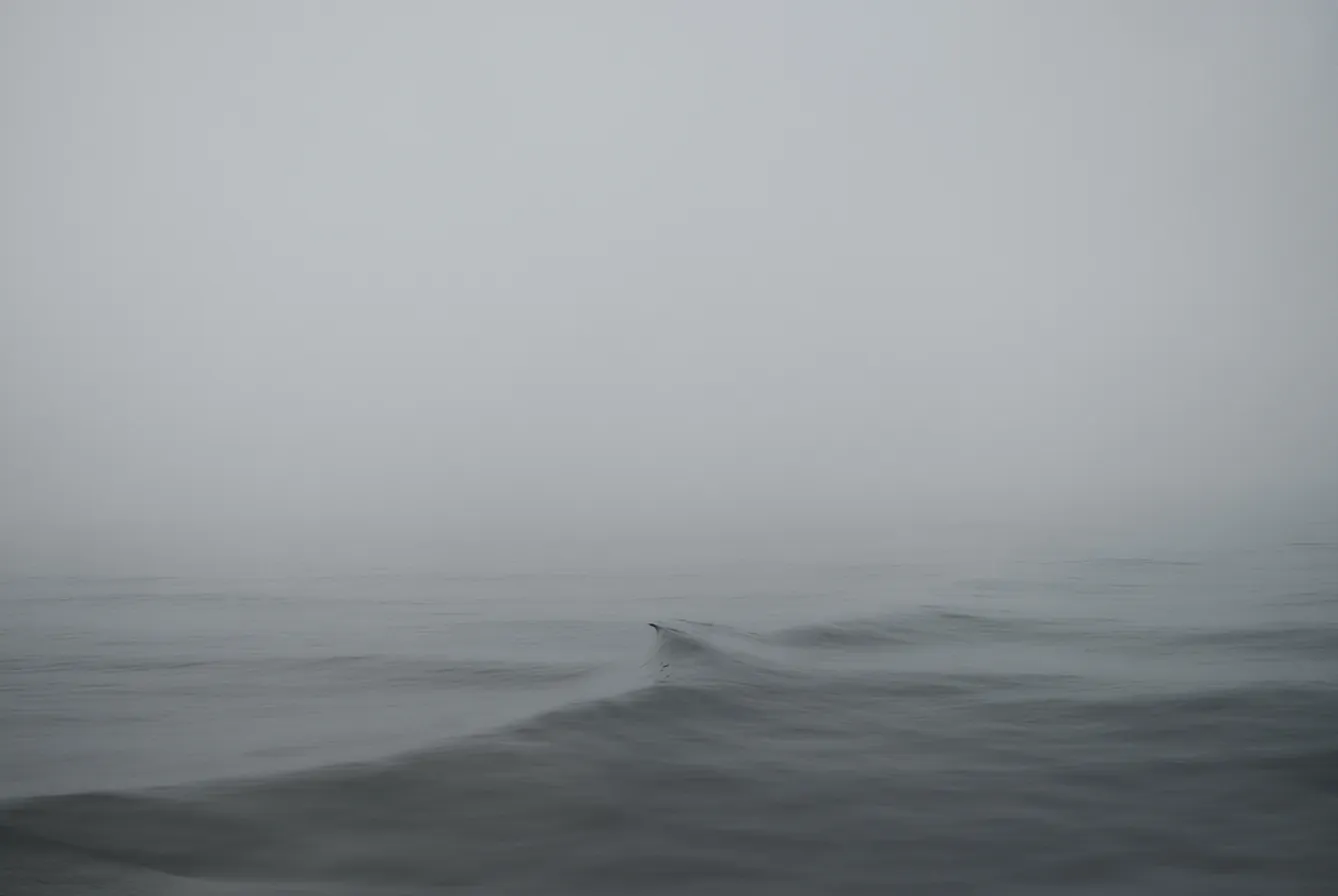 A small wave in a grey, misty sea with a barely visible horizon, photographed by Stephan Vanfleteren.
