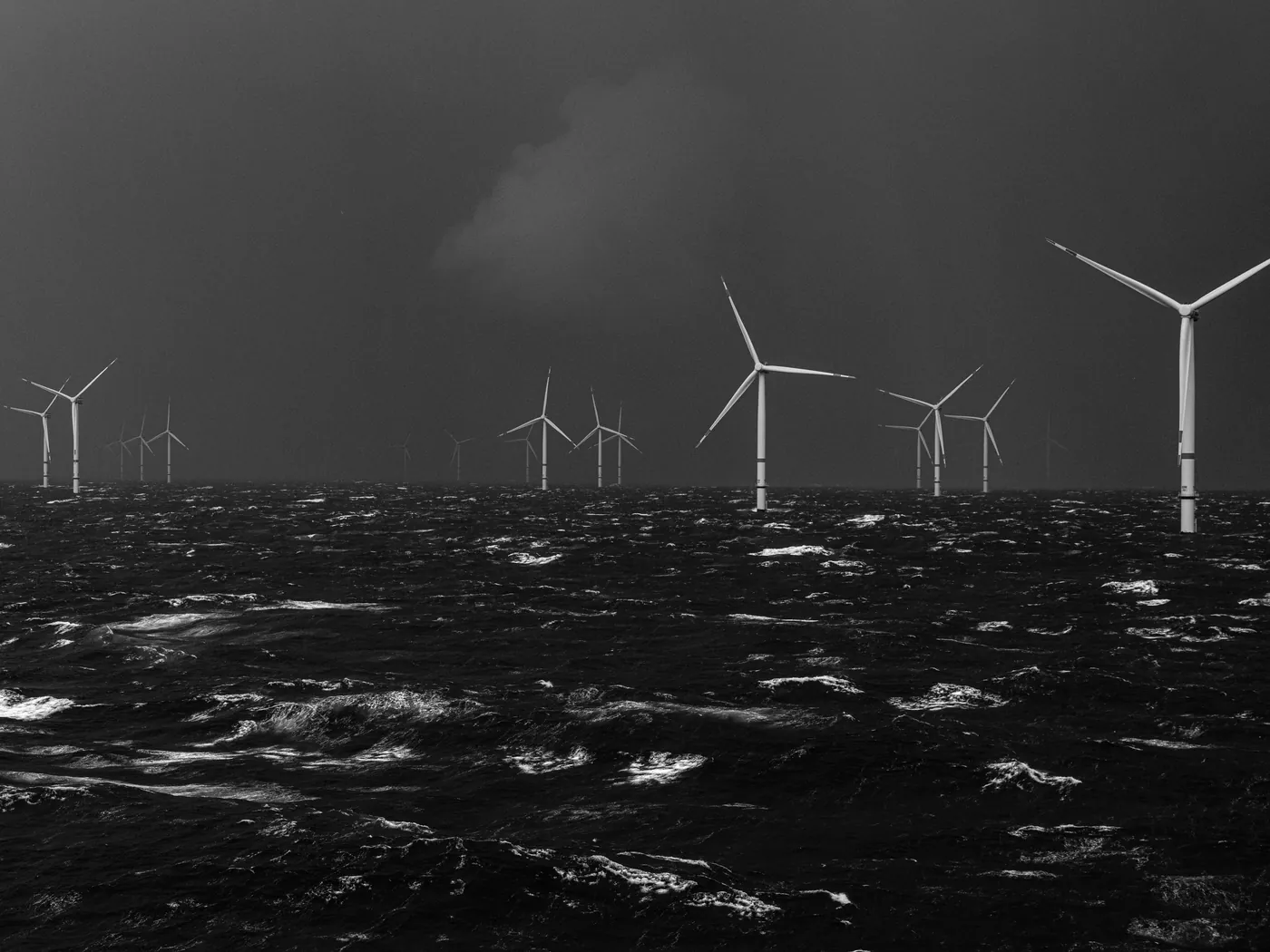 Zwart-witfoto van windturbines op een woelige zee onder donkere wolken, door Kadir van Lohuizen.