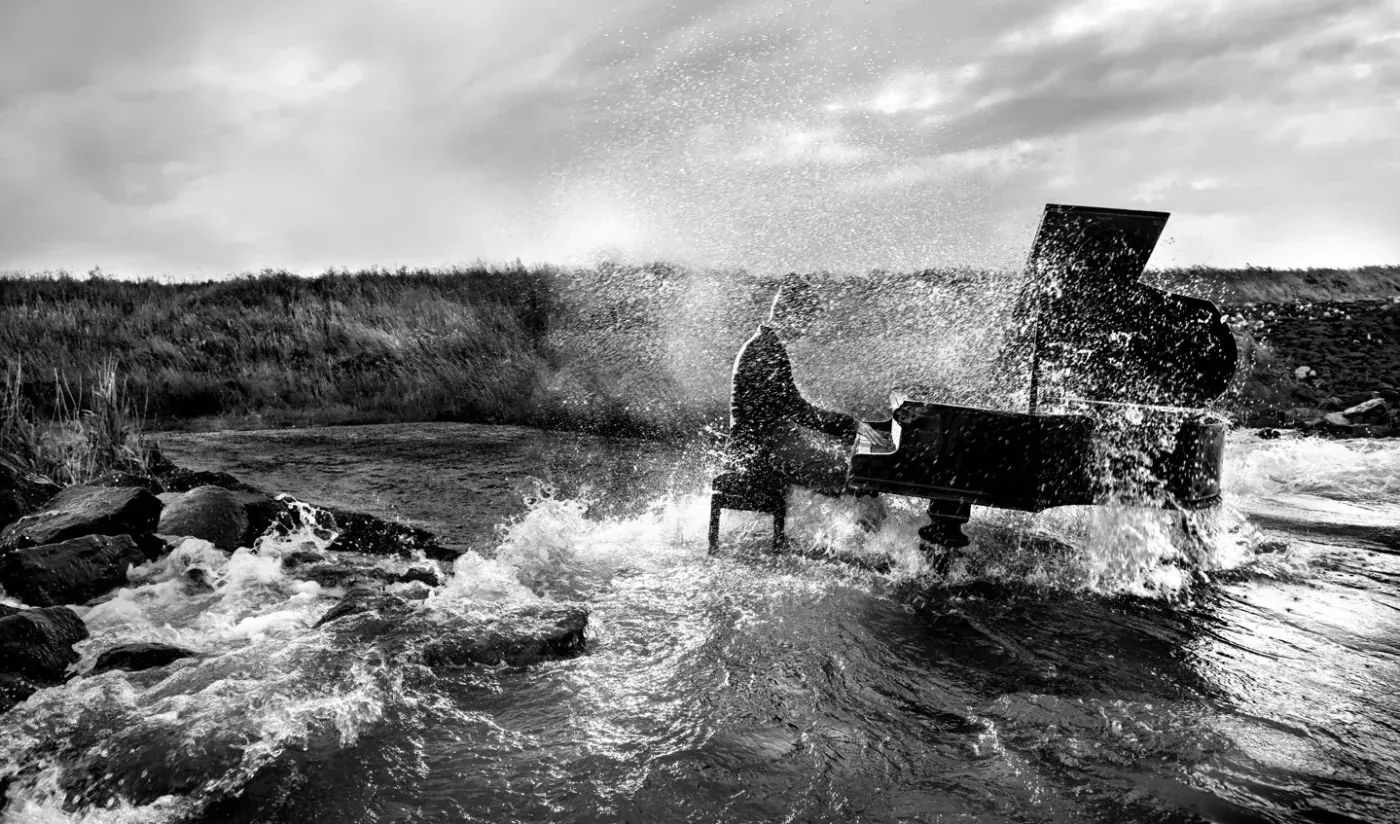 Black-and-white image of a pianist at a grand piano among rocks and splashing water.