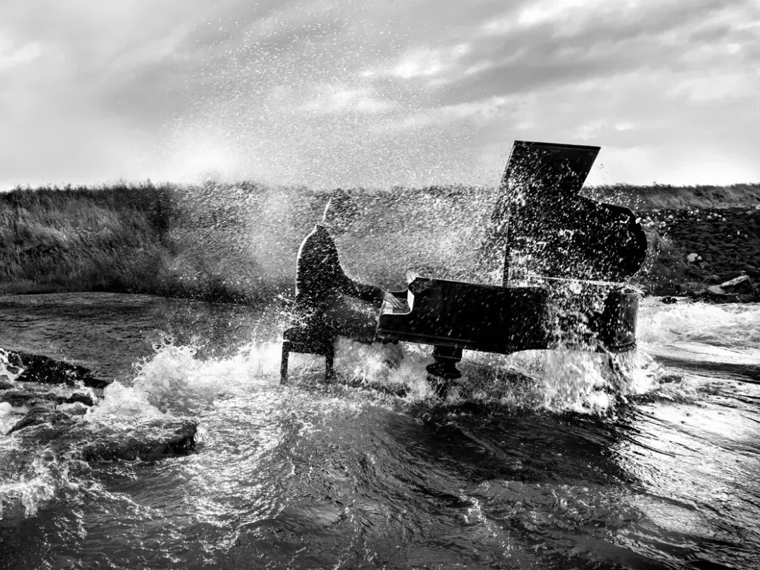 Zwart-witbeeld van een pianist aan een vleugel tussen rotsen en opspattend water.