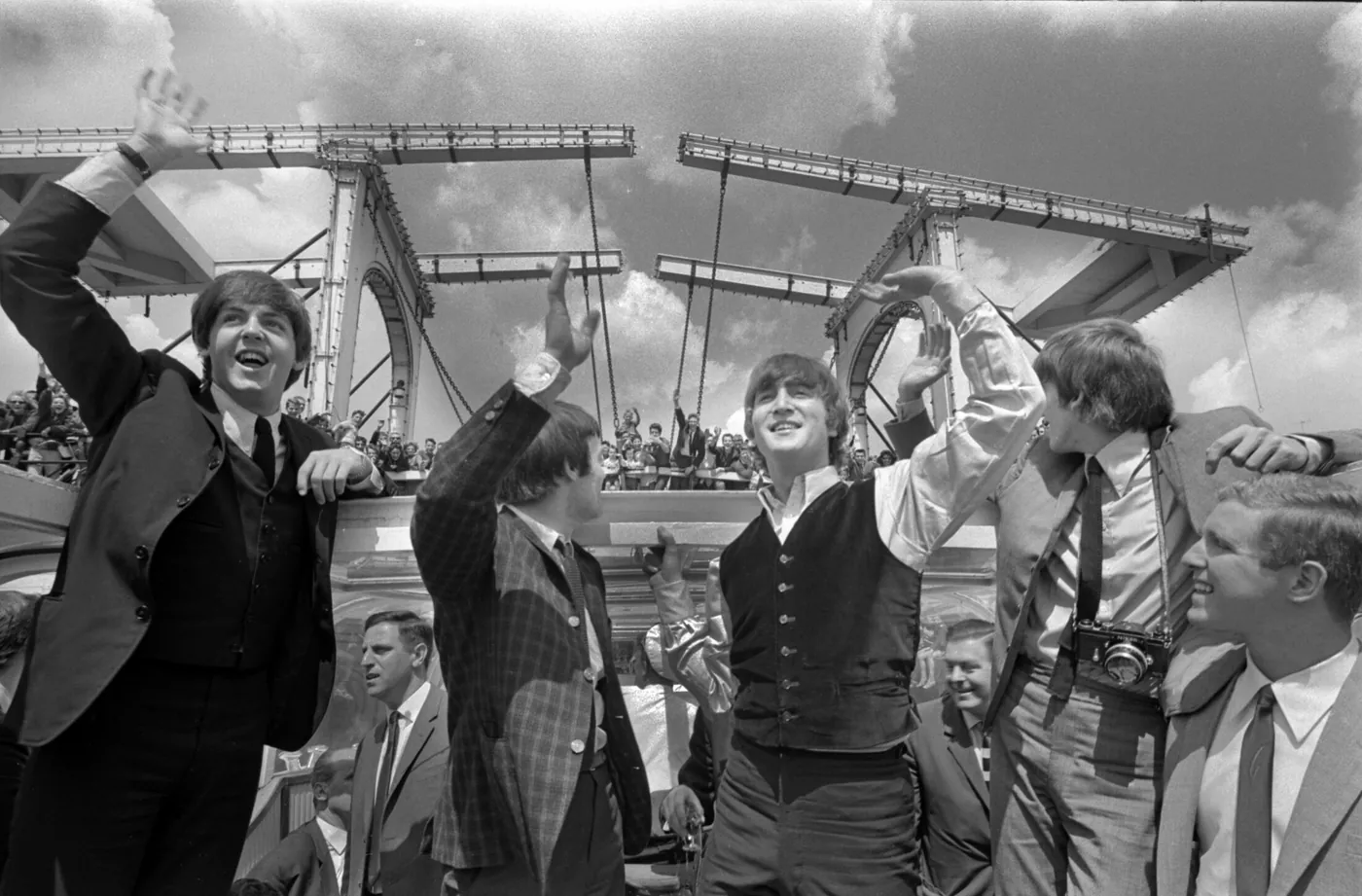 Zwart-witfoto van The Beatles die vanaf een rondvaartboot zwaaien, met een ophaalbrug en publiek op de achtergrond.
