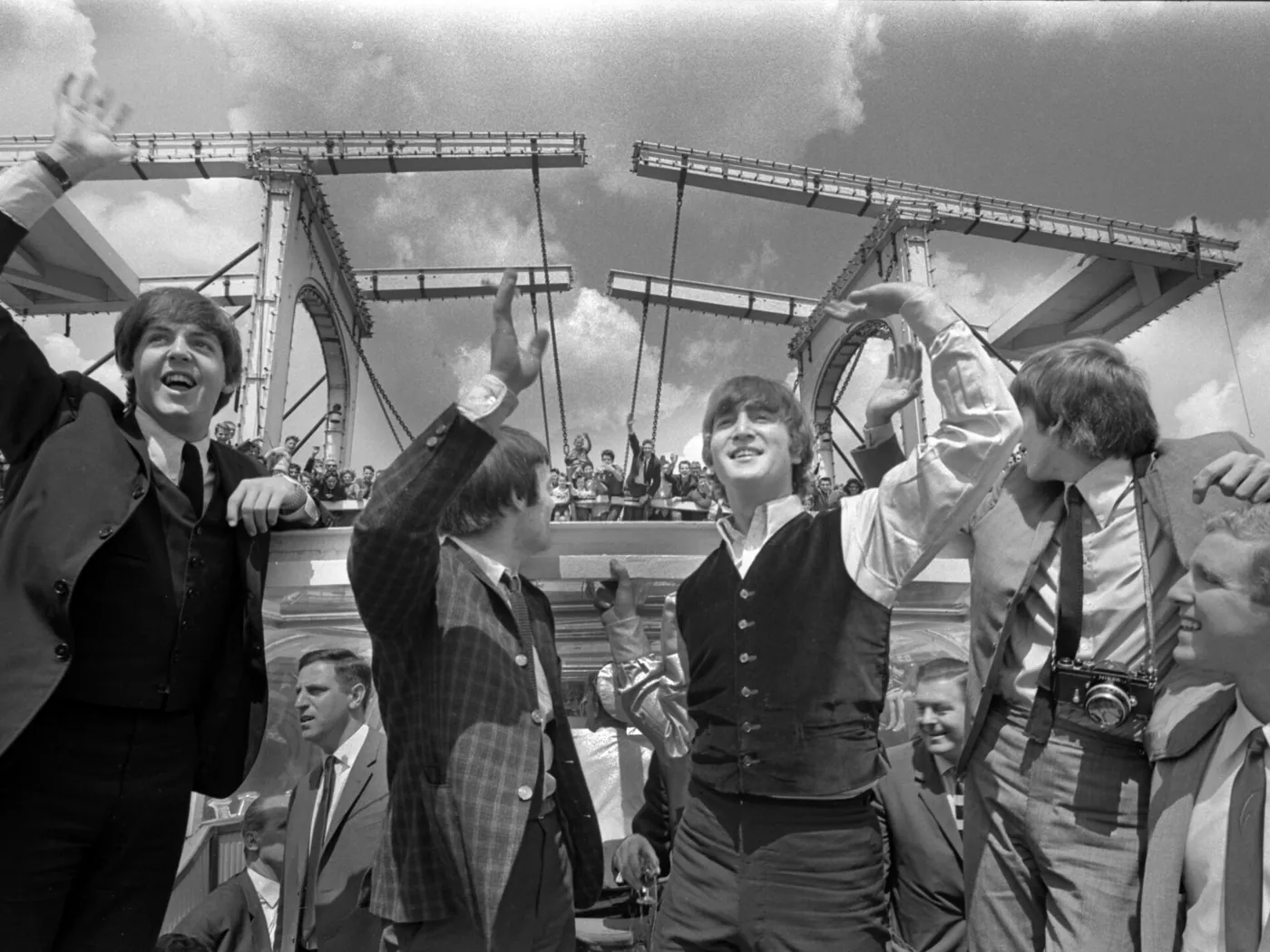 Zwart-witfoto van The Beatles die vanaf een rondvaartboot zwaaien, met een ophaalbrug en publiek op de achtergrond.