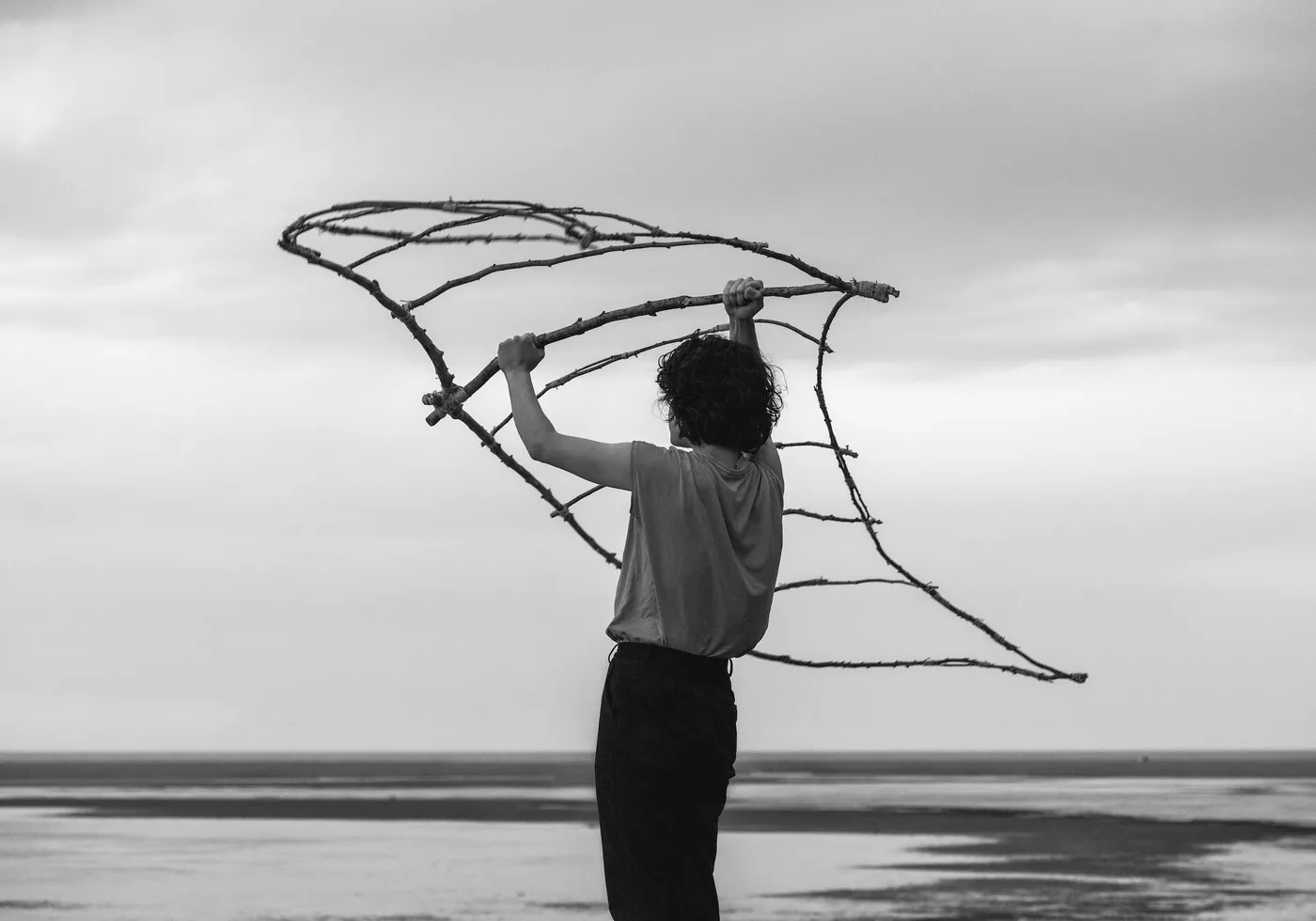 Black-and-white photograph of a person holding a structure of branches overhead by the water, by Chloé Azzopardi.
