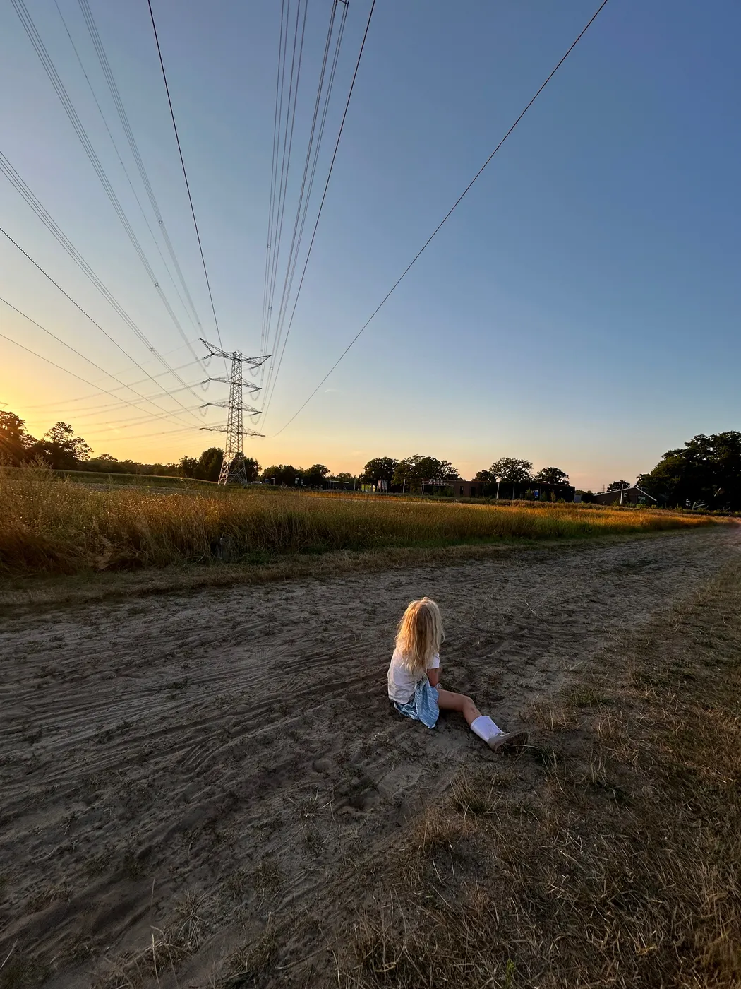 Girl sitting on a sandy path beneath power lines at sunset, photographed by Bettina Hogeweg for Alphameisje