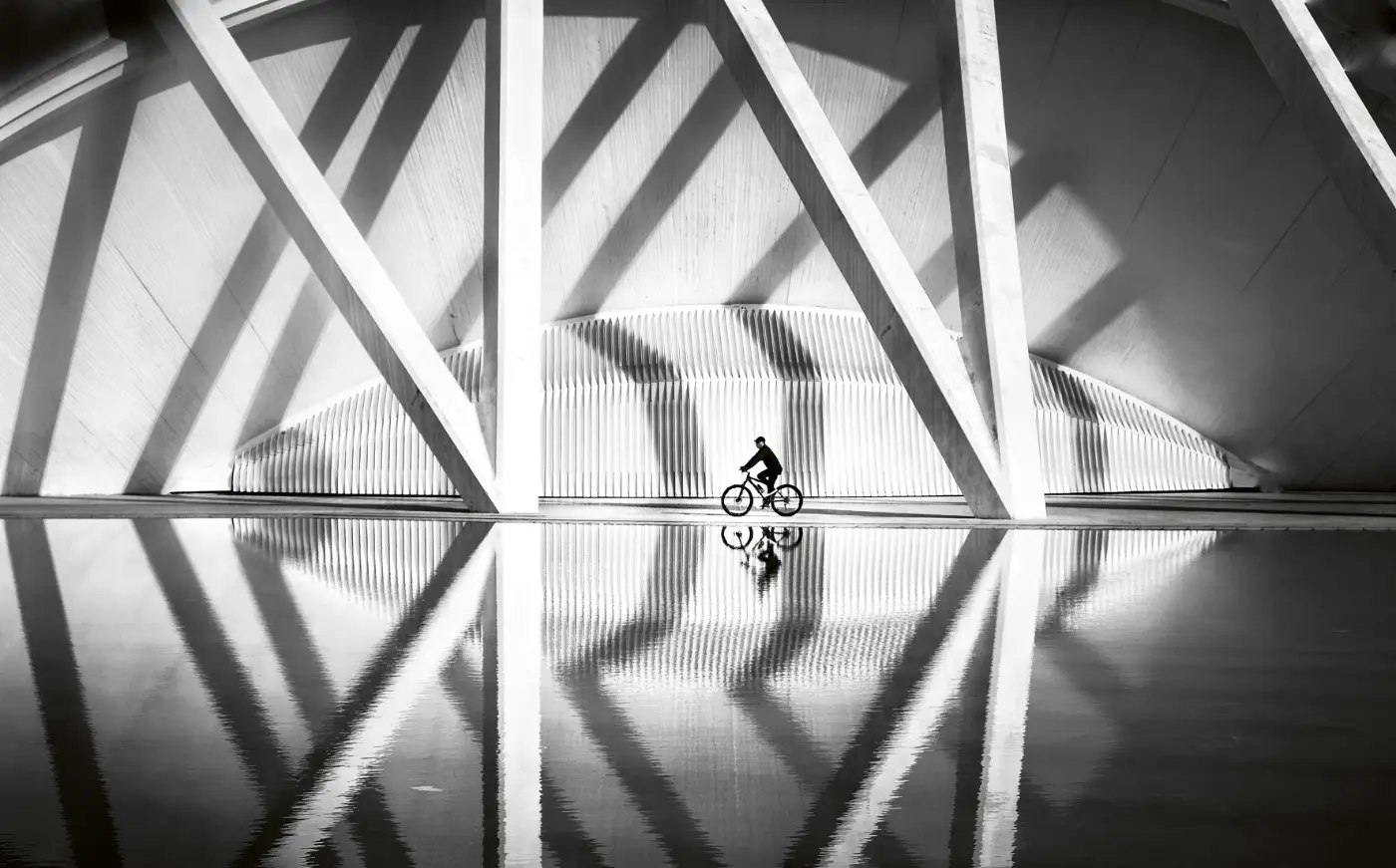 Zwart-witfoto van een fietser langs geometrische architectuur, weerspiegeld in het water