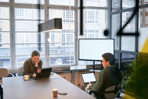 Two men working on laptops in a conference meeting room at betahaus coworking, with large windows and a monitor on the side.