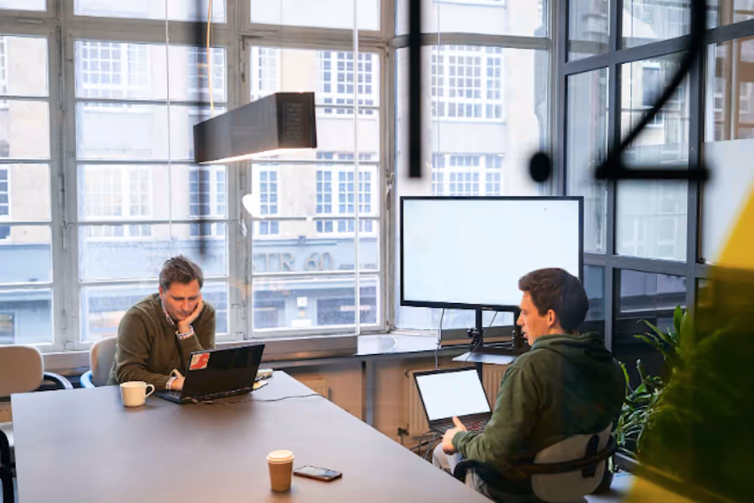 Two men working on laptops in a conference meeting room at betahaus coworking, with large windows and a monitor on the side.