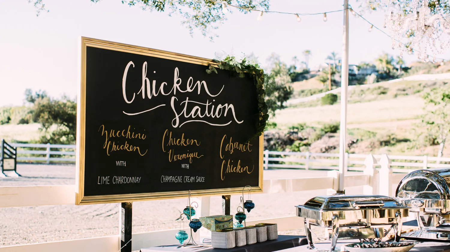 Outdoor buffet setup with a black chalkboard menu titled 'Chicken Station' listing Zucchini Chicken with Lime Chardonnay, Chicken Veronique with Champagne Cream Sauce, and Cabernet Chicken.