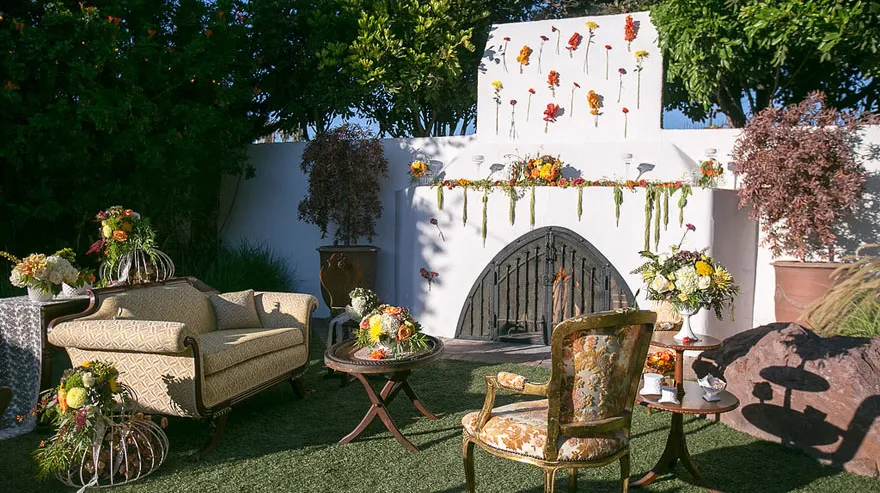 Outdoor seating area with vintage furniture and floral arrangements in front of a white fireplace wall decorated with hanging flowers.