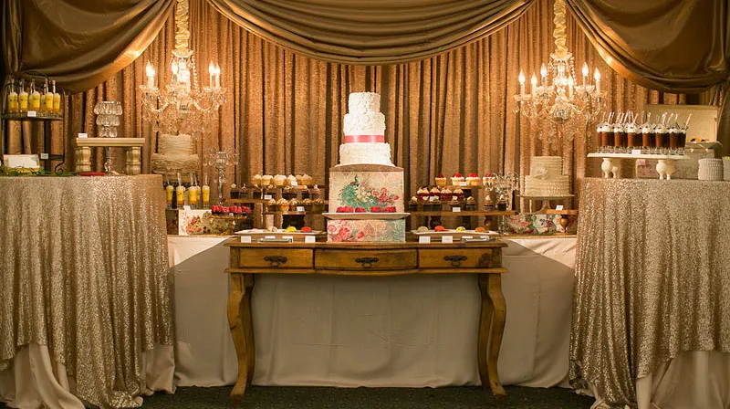 Elegant dessert table with a three-tiered decorated cake and assorted cupcakes under chandeliers and gold draped curtains.