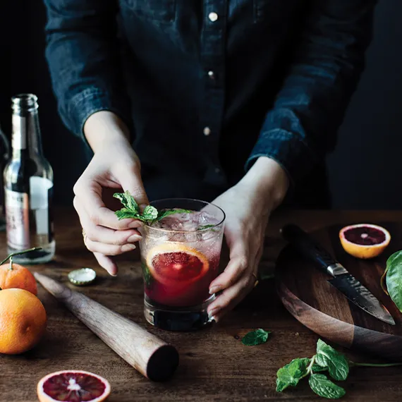 Person garnishing a blood orange cocktail with fresh mint on a wooden table with citrus fruits, muddler, and knife.