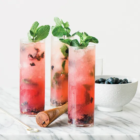Three tall glasses of pink mint and berry cocktails on a marble surface with a wooden muddler and a bowl of blueberries.
