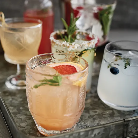 Variety of colorful cocktails garnished with fruit and herbs on a metal tray.
