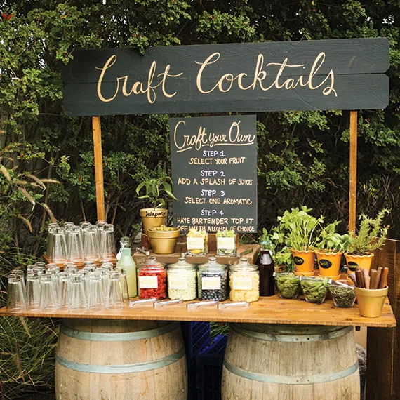 Outdoor craft cocktail bar with jars of fruit, herbs, spices, glassware, and a chalkboard showing steps to craft your own cocktail.