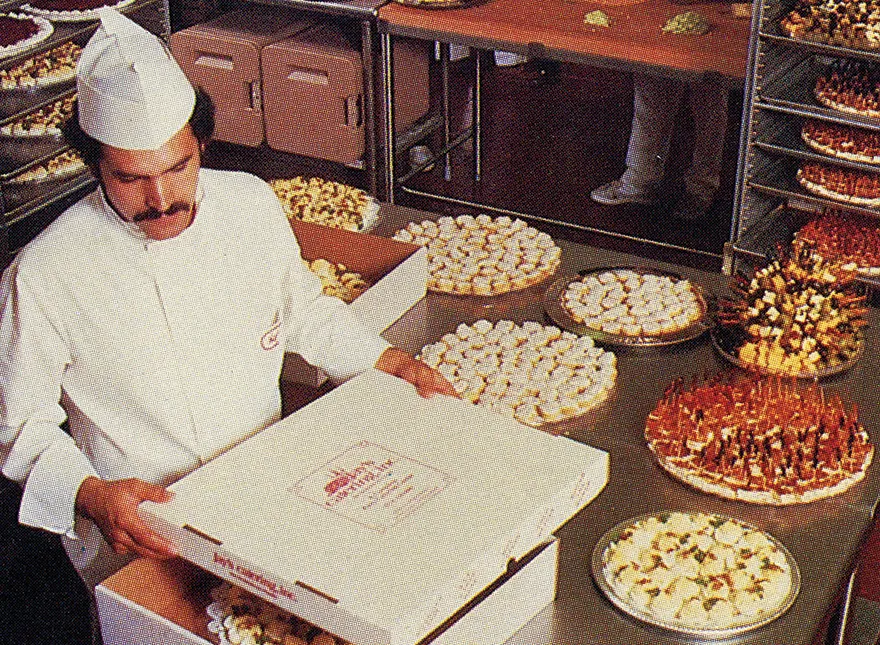 Man in white baker uniform and hat packing assorted pastries arranged on trays in a bakery kitchen.