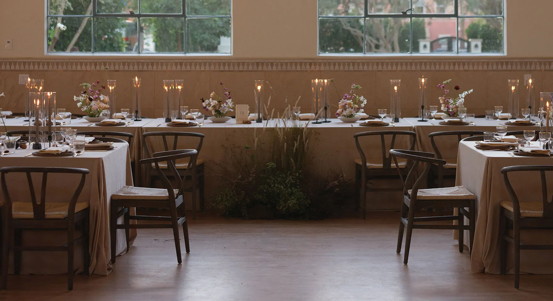 Elegantly set banquet tables with beige tablecloths, wooden chairs, floral centerpieces, and tall candle holders inside a room with large windows.