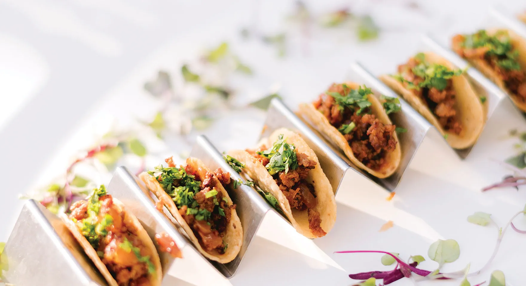Row of mini tacos filled with seasoned meat and topped with chopped cilantro, served in a stainless steel taco holder on a white surface with scattered greens.