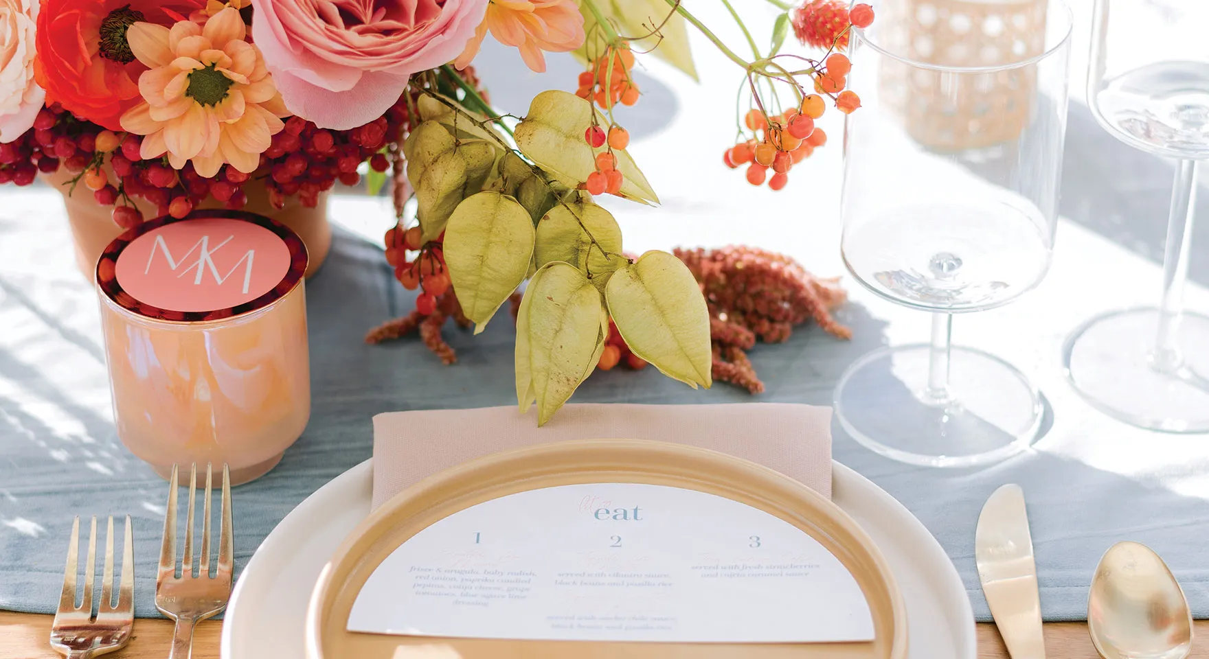 Table setting with gold-rimmed plate, menu card, gold cutlery, wine glasses, peach candle, and floral centerpiece with pink and red flowers and orange berries.