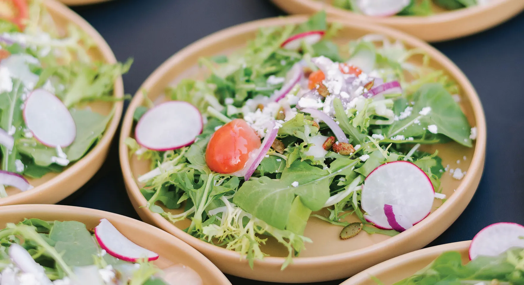 Fresh mixed green salad with radish slices, cherry tomatoes, red onion, pumpkin seeds, and crumbled cheese on a beige plate.