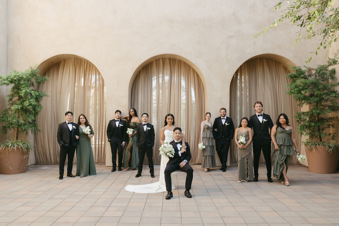 Wedding party poses for group photo in front of 3 stucco archways draped in taupe fabric. The bridesmaids are dressed in shades of green, the groomsmen and groom are dressed in black, and the bride wears white.