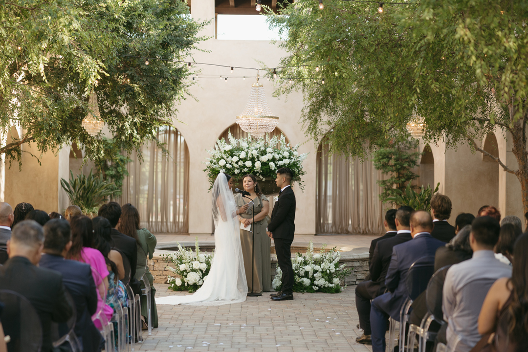 Looking down the aisle at the bride and groom reading handwritten vows as guests sit in ghost chairs and draped archways and Spanish Fountain add to the ambiance.