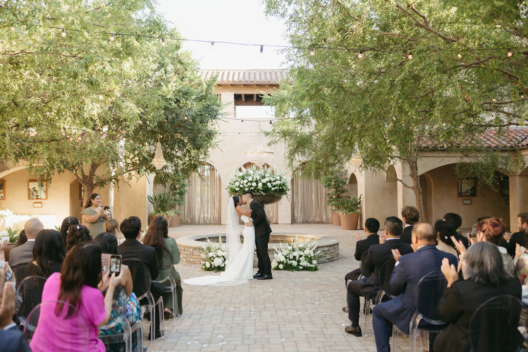 Bride and groom share their first kiss under a canopy of trees in the Serra Plaza Courtyard surrounded by the venue's European architecture.