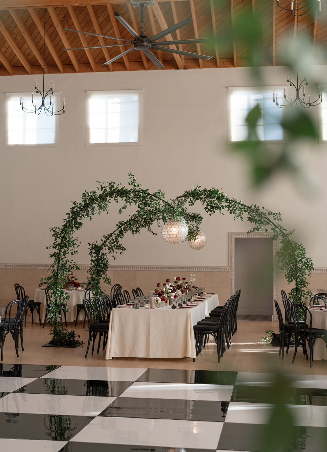 Greenery arches with pendant lights hang above a reception table topped with red wine-colored napkins and floral centerpieces.