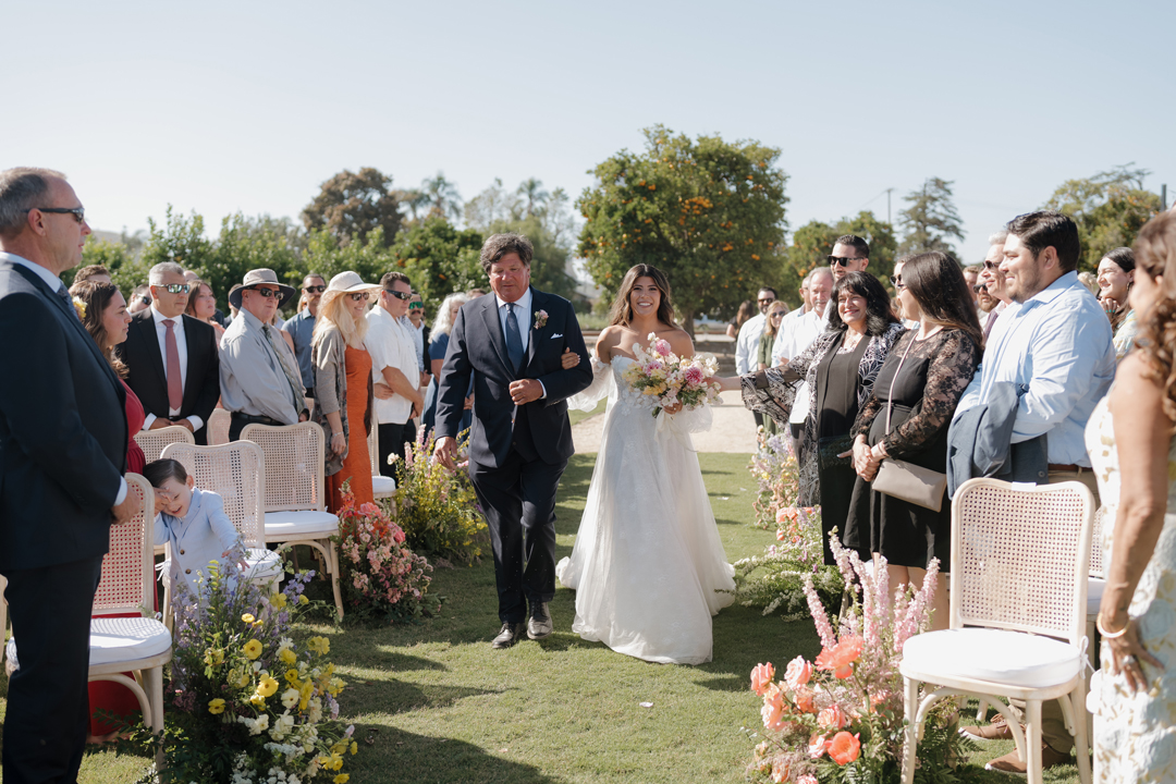 Bride and dad walk arm in arm into outdoor wedding ceremony on grassy field.