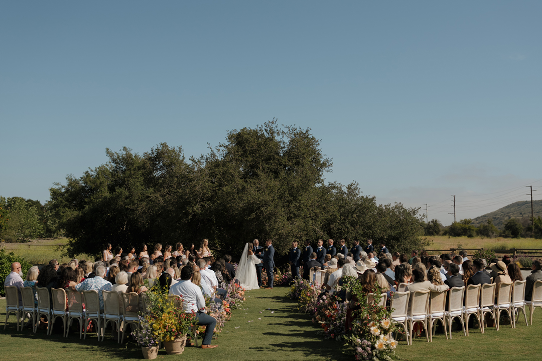 Outdoor wedding ceremony on grassy field with colorful flowers.