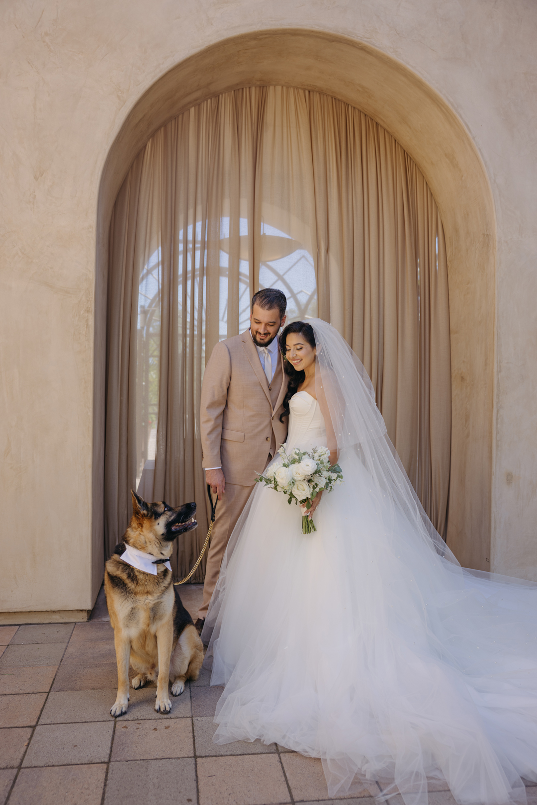 Bride and groom smile at dog in front of Serra Plaza's large draped archway in the Courtyard.
