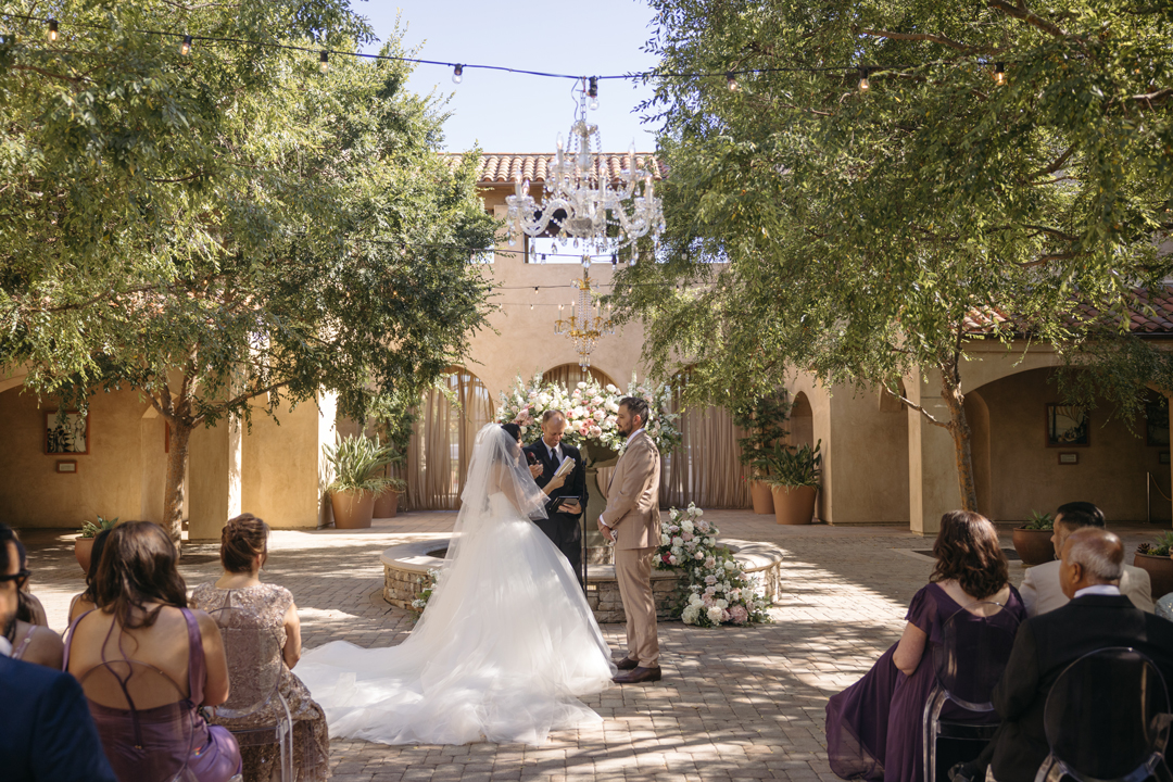 Bride and groom read handwritten vows in front of draped archways and Spanish fountain.