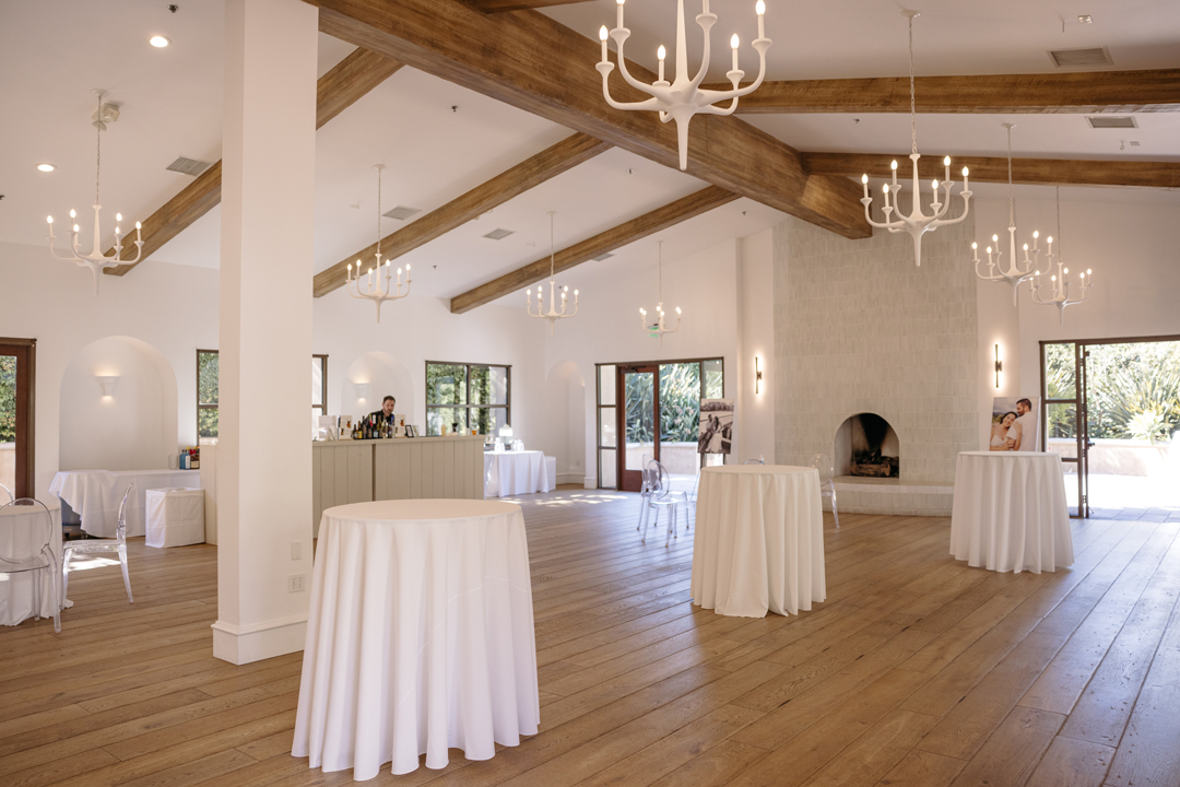 Wedding cocktail hour lounge set with white draped tables inside Spanish Revival wedding venue.