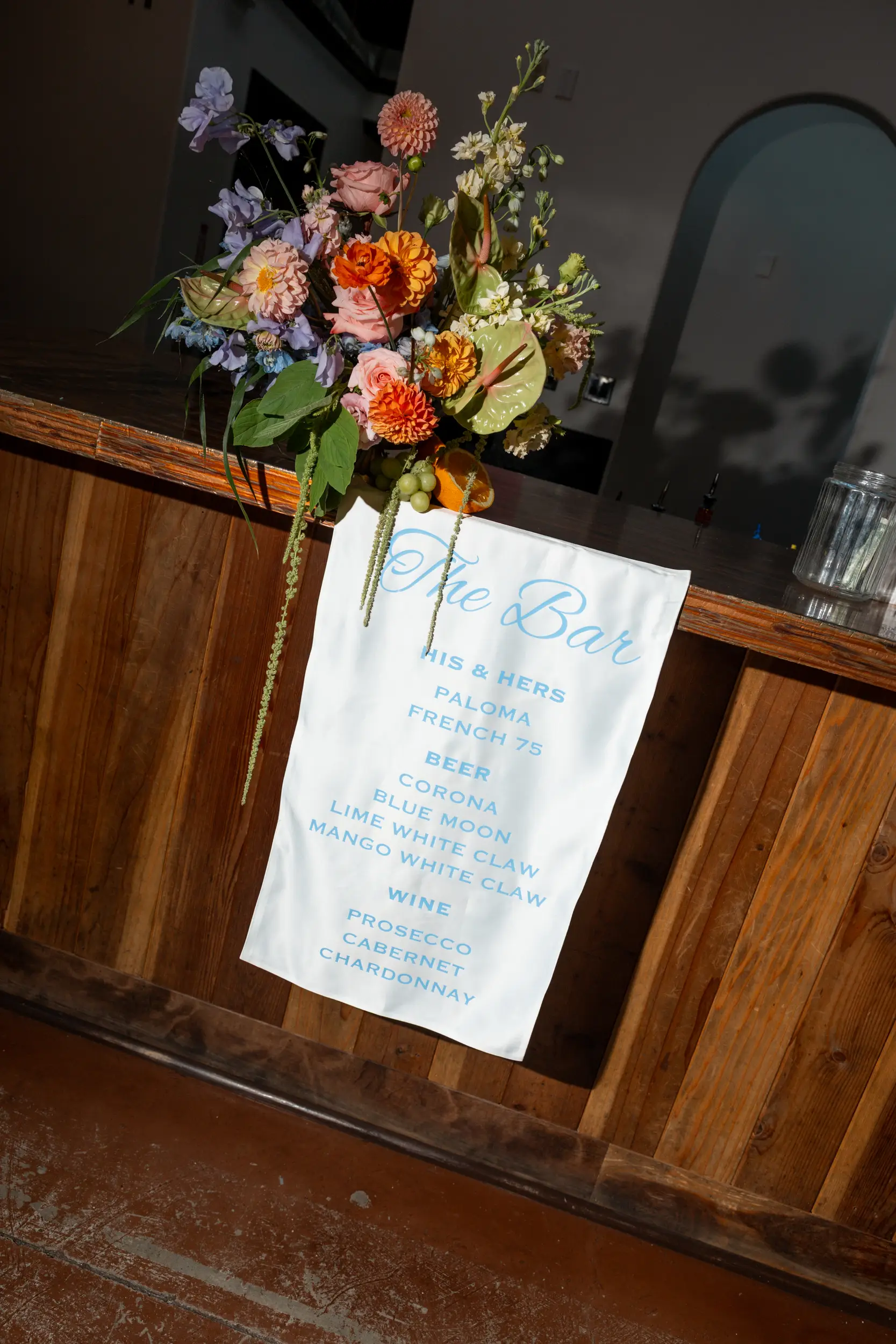 Colorful florals are placed atop the bar and a fabric bar sign drapes over the edge with the names of each signature drink.