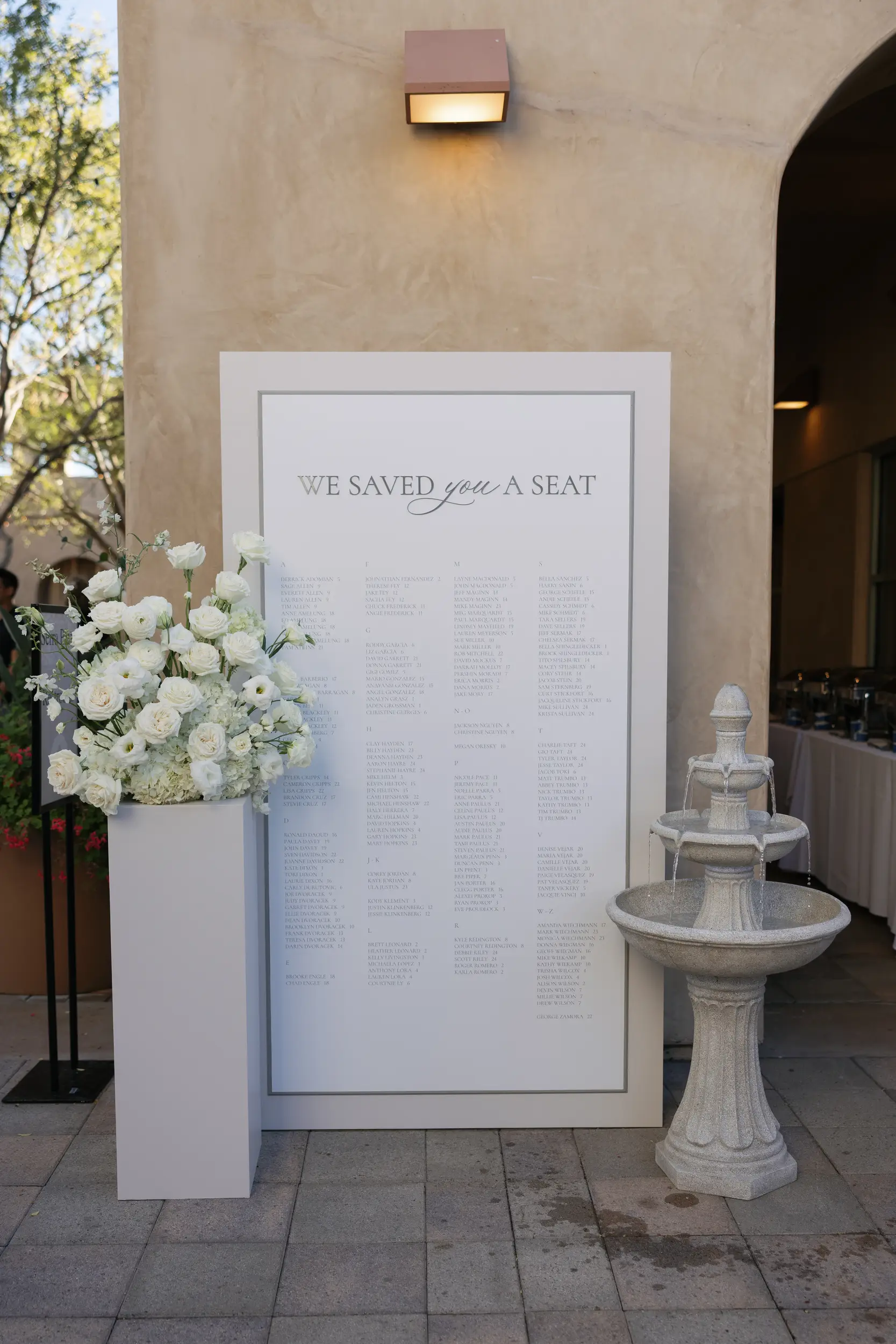 Romantic wedding seating chart with white florals, neutral text, and fountain.