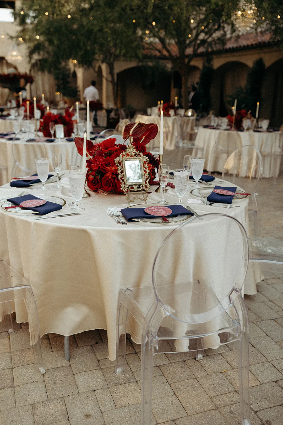 Reception table with a red floral centerpiece, navy napkins and clear chairs.