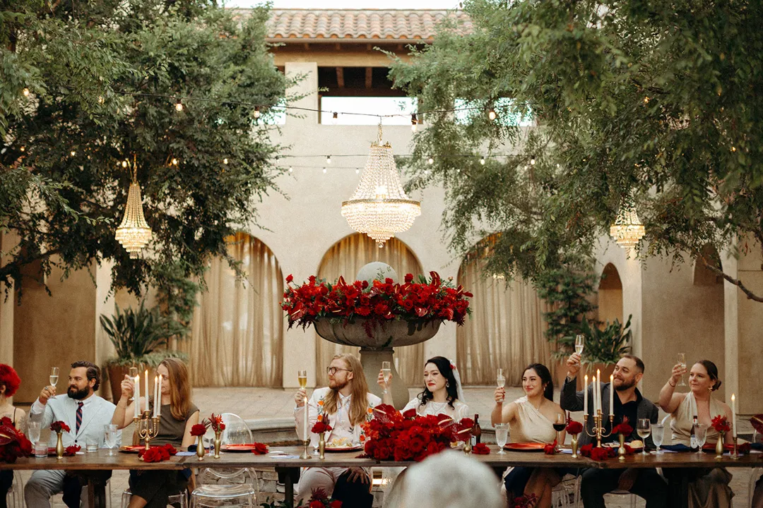 Wedding party seated at a courtyard reception table beneath chandeliers.